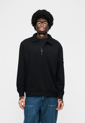 Mlada oseba z vitiligom, oblečena v črn pulover z zadrgo in modre kavbojke, stoji pred enostavnim belim ozadjem.