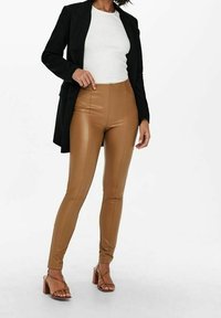 Hnědé legíny ze syntetické kůže s vysokým pasem v kombinaci s přiléhavým bílým topem a černým sako; celý outfit doplňují hnědé sandály na vysokém podpatku.