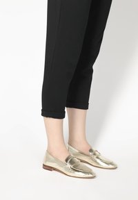 Goldene metallische Slipper mit rundem Zehenbereich und dekorativem Riemen, kombiniert mit schwarzen cropped Pants. Glatte Textur und dezenter Glanz.