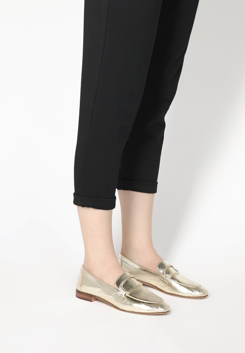 Goldene metallische Slipper mit rundem Zehenbereich und dekorativem Riemen, kombiniert mit schwarzen cropped Pants. Glatte Textur und dezenter Glanz.