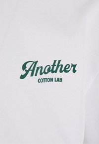 Balta medvilninė marškinėliai su žaliais užrašais "Another" virš "COTTON LAB", užrašyti klasikinio stiliaus šriftu ir minkšta tekstūra.