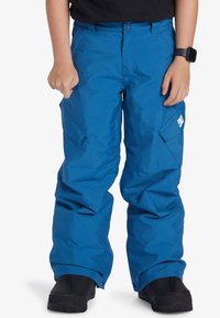 Blå snöbyxor gjorda av vattentätt material. Funktioner inkluderar cargofickor, avspänt snitt och förstärkta fållar. Svarta skor synliga längst ner.