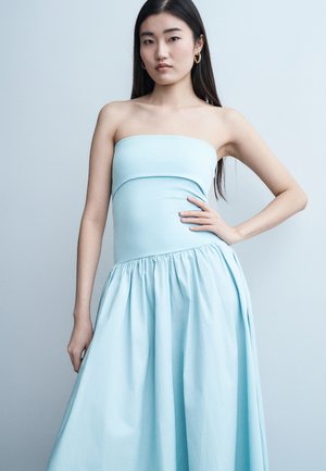 Νεαρή γυναίκα με μακριά σκούρα μαλλιά, ντυμένη με ένα αμάνικο ανοιχτό μπλε φόρεμα, που στέκεται με το ένα χέρι στη μέση της μπροστά σε ένα ανοιχτό μπλε φόντο.