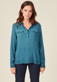 Maglietta a maniche lunghe color teal con scollo a V, due taschini sul petto e orlo leggermente curvo. Tessuto morbido e liscio con cuciture minime.