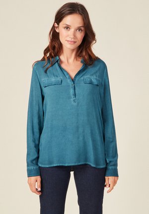 Camisa de manga larga color teal con escote en V, dos bolsillos en el pecho y un bajo ligeramente curvado. Textura suave y lisa con costuras mínimas.
