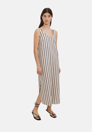 Mujer con un vestido beige sin mangas con rayas negras verticales y sandalias negras planas con tiras, de pie frente a un fondo liso.