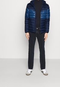 Marineblauw en blauwe donsjas met capuchon, met horizontale strepen, gedragen over een zwarte coltrui en gecombineerd met donkere jeans en witte sneakers.