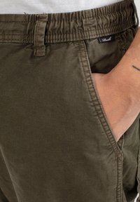 Hand in pocket of olive green elastic waist pants with belt loop and small black fabric brand tag.