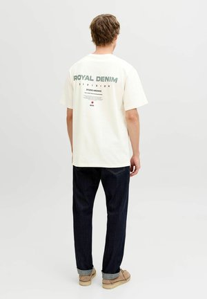 Moški s svetlo rjavimi lasmi, oblečen v belo srajco s kratkimi rokavi z napisom "ROYAL DENIM", temno modre kavbojke in bež čevlje, stoji z obrnjenim hrbtom.