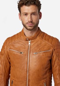 Bruine lederen motorjack met gequiltte schouderaccenten, twee borstzakken met rits en een hoge kraag, met zilverkleurige hardware en een gladde textuur.