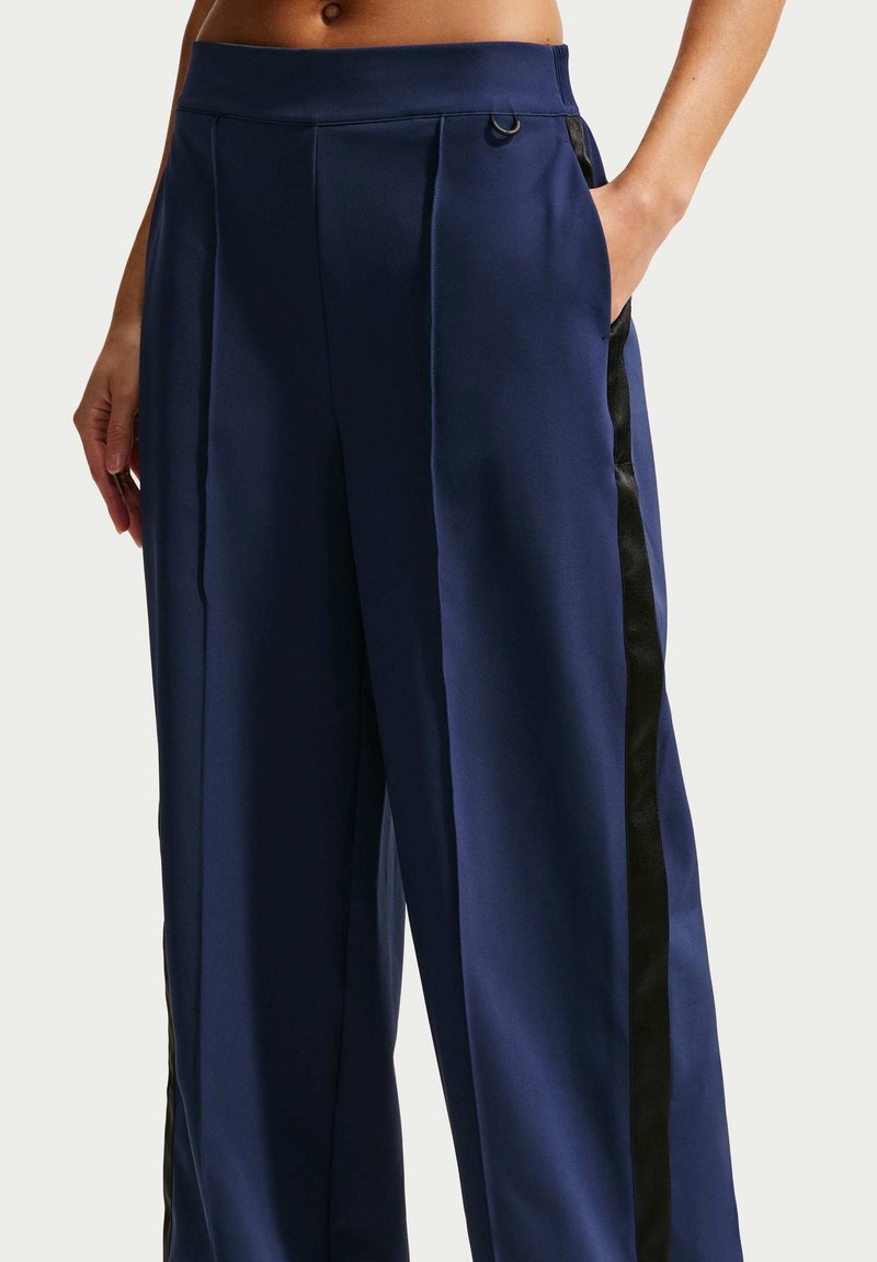 Person trägt marineblaue, hoch taillierte, weit geschnittene Hosen mit schwarzen Seitenstreifen, eine Hand in der Tasche, sichtbarer bauchfreier Oberkörper.