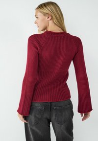 Roter gerippter Strickpullover mit Rundhalsausschnitt und ausladenden Ärmeln, kombiniert mit dunkelgrauen Jeans. Strukturiertes Gewebe mit vertikalen Linien.