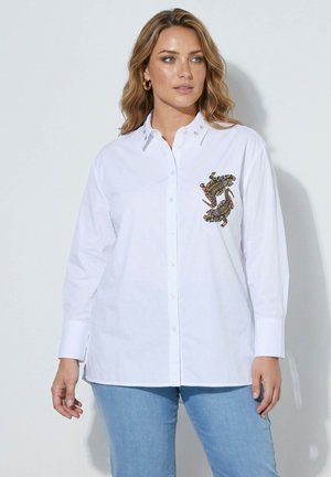 Mujer que lleva una camisa blanca con botones y ranas bordadas en el lado izquierdo del pecho, combinada con vaqueros azul claro, de pie frente a un fondo liso.