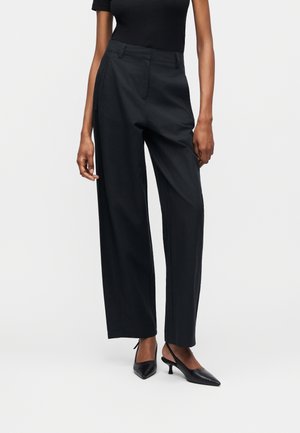 Žena oblečená v čiernych širokých nohaviciach s vysokým pásom a čiernych sandáloch na nízkom podpätku so zadným remienkom, stojaca pred jednofarebným svetlým pozadím.