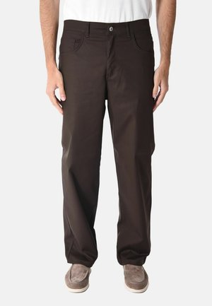 Uomo che indossa pantaloni larghi marrone scuro, camicia bianca e scarpe slip-on in pelle scamosciata marrone chiaro, in piedi con le mani rilassate lungo i fianchi.
