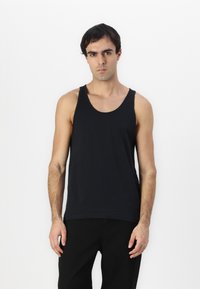 Débardeur en coton noir avec un col rond et de larges emmanchures, associé à un pantalon noir, porté par un mannequin sur un fond blanc uni.