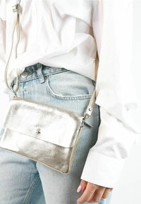 Henkilö, jolla on päällään vaaleansiniset farkut ja valkoinen pitkähihainen paita, kantaa metallinhohtoista hopeista olkalaukkua, jossa on tähden yksityiskohta.