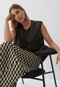 Top nero senza maniche con zip frontale, abbinato a una gonna a fantasia con forme geometriche in toni scuri e chiari. Seduta su una sedia nera.