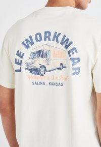 Kremo spalvos marškiniai su iliustracija apie pristatymo mikroautobusą, ant kurio užrašyta "LEE WORKWEAR" ir šūkis "Darbo drabužiai yra mūsų siela" mėlyna ir oranžine spalvomis.