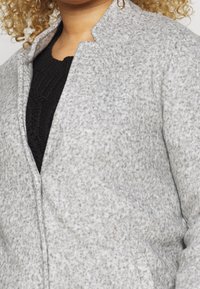 Grau strukturierte Jacke mit einem kragenlosen Design, ausgestattet mit einem Frontreißverschluss und seitlichen Taschen. Getragen über einem schwarzen Strickpullover.