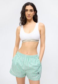 Νεαρή γυναίκα με σγουρά μαλλιά, που φοράει λευκό αθλητικό σουτιέν και πράσινο-λευκές ριγέ βερμούδες, στέκεται με τα χέρια στις τσέπες.