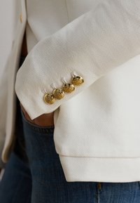Hvid struktureret blazerærme med fire dekorative gyldne knapper over blå denim jeans.