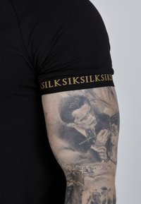 Crna košulja kratkih rukava s teksturiranom zlatnom logotip trakom na gornjem dijelu ruke, nosi se preko detaljno tetovirane podlaktice.