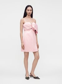 Femme portant une mini-robe rose sans manches avec un grand nœud sur le devant et des escarpins slingback noirs à bout pointu.
