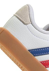 Zapatilla blanca de cuero con un acabado texturizado, que presenta franjas azules y rojas, suela de goma y un detalle en gamuza en el talón.