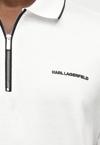 Polo blanc avec une fermeture éclair noire et le logo noir "KARL LAGERFELD" sur la poitrine, porté par une personne à la peau foncée.