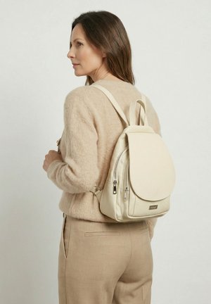 Žena oblečená v béžovém svetru a kalhotách, nesoucí na zádech světle krémový kožený batoh se dvěma kapsami na zip.