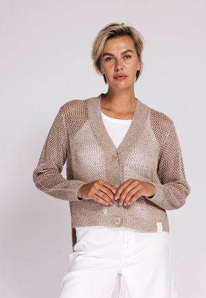 Frau mit kurzen blonden Haaren, die eine beige offene Strickjacke mit großen Knöpfen über einem weißen Top und weißen Hosen trägt, steht vor einem einfarbigen Hintergrund.