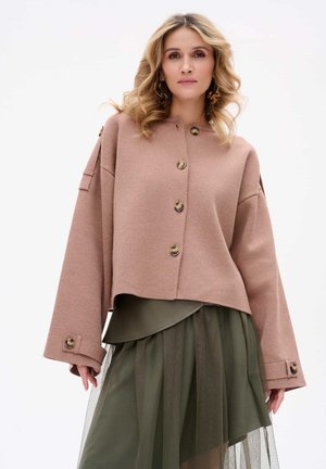 Pink cropped sweater with large buttons and wide sleeves, layered over an olive green dress with a translucent skirt and side accents.