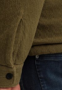 Zbliżenie na rękaw oliwkowozielonej kurtki o teksturze z czarnym guzikiem i kieszenią z ciemnoniebieskich dżinsów.