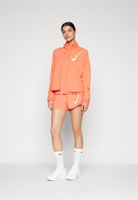Veste de sport orange vif avec un col haut, shorts assortis et logos Nike blancs. Portée avec des chaussettes et des chaussures blanches. Tissu lisse.
