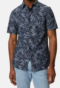 Mann in einem kurzärmligen, knöpfbaren Hemd mit dunkelblauem tropischen Blättermuster auf grauem Stoff, kombiniert mit hellblauen Jeans.