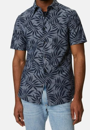 Mann in einem kurzärmligen, knöpfbaren Hemd mit dunkelblauem tropischen Blättermuster auf grauem Stoff, kombiniert mit hellblauen Jeans.