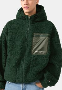 Hustá tmavozelená fleecová bunda so zipsovou olivovozelenou vreckom, kapucňou a nastaviteľnými šnúrkami. Na vrecku je umiestnené logo.