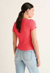 Kvinde med mørkt hår i hestehale iført en krøllet, lyserød kortærmet top og lyseblå højtaljede jeans, vendt væk.