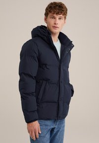 Navy puffer jacket with a hood, featuring a zip and button closure, textured fabric, and side pockets, worn over a light blue shirt.