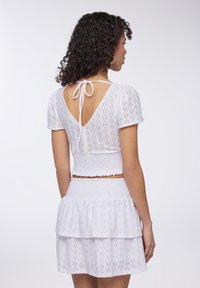 Crop top bianco arricciato e gonna a strati, con motivi a occhio di pernice e retro a V con lacci, maniche corte a sbuffo e tessuto testurizzato.