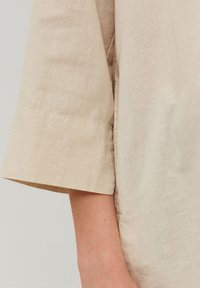 Beige linnedragt med bred, foldet kortærme. Tekstureret stof og rent, minimalistisk design. Søm detalje langs siden.