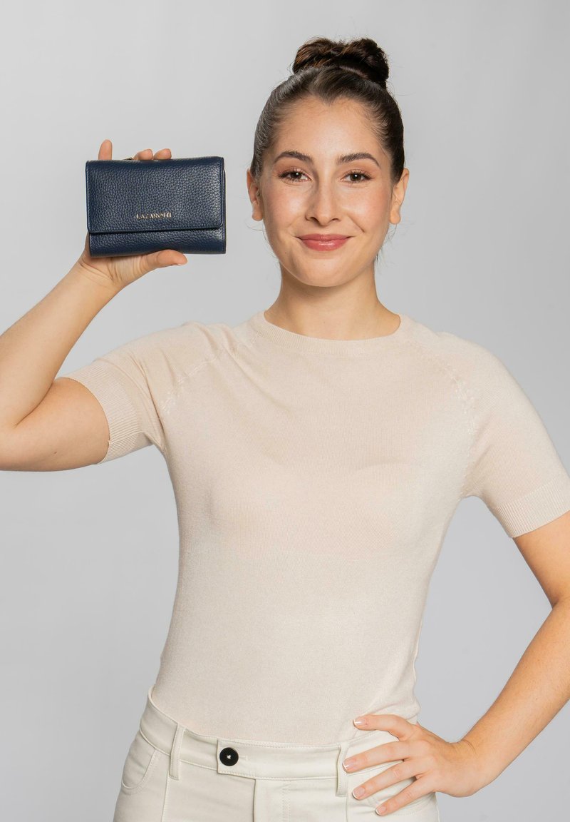 Billetera de piel azul marino con acabado texturizado, que presenta un cierre de solapa y el logo de la marca, sostenida por una persona contra un fondo neutro.