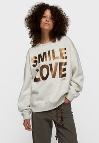 Sudadera gris claro con texto met�álico dorado "SMILE" y "LOVE". Mangas anchas y ajuste relajado. Diseño casual con detalles visibles de costura.