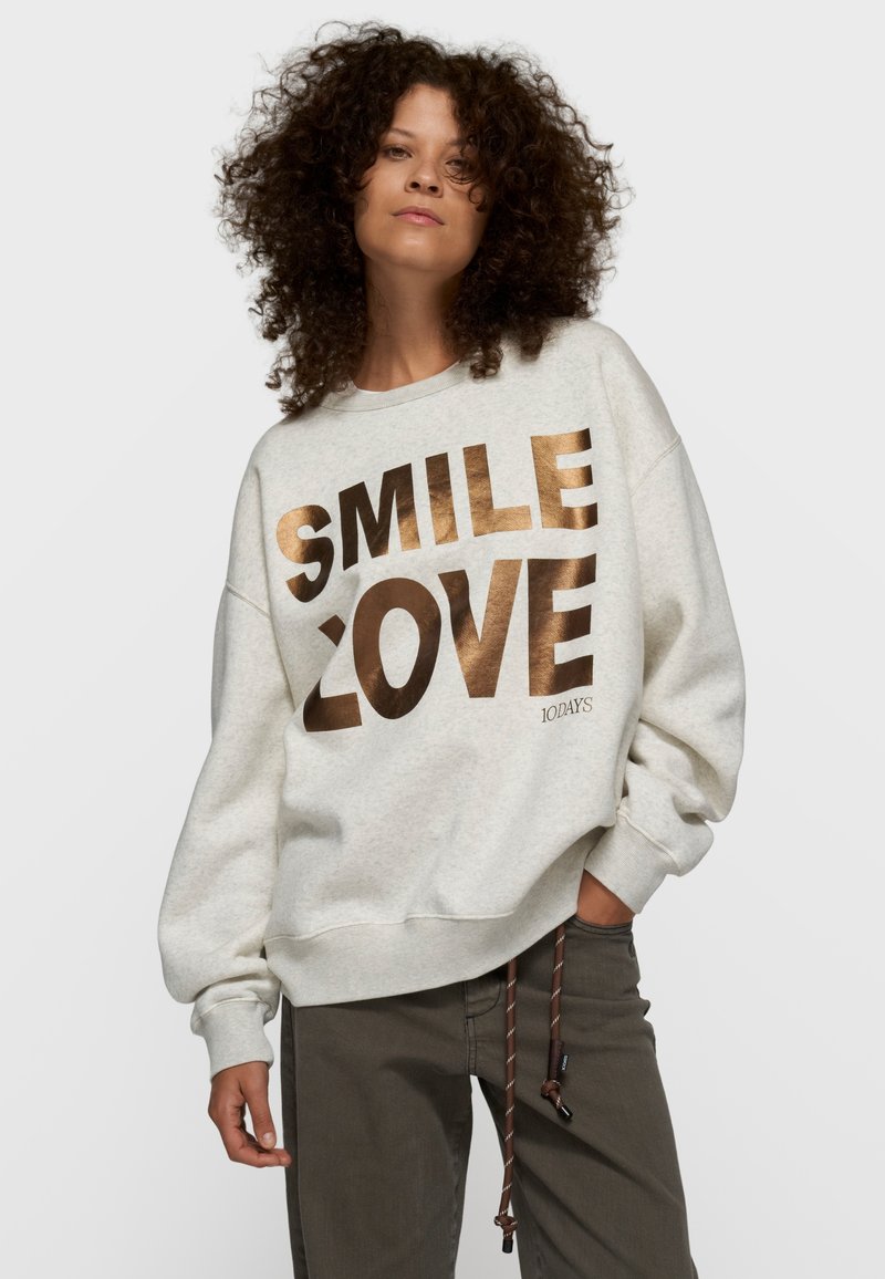 Sudadera gris claro con texto metálico dorado "SMILE" y "LOVE". Mangas anchas y ajuste relajado. Diseño casual con detalles visibles de costura.