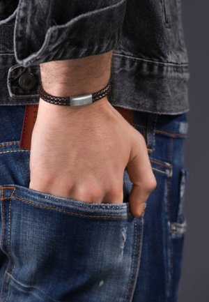 Braccialetto intrecciato in pelle marrone con un elemento metallico rettangolare argentato, indossato al polso, con la mano appoggiata in una tasca di denim.
