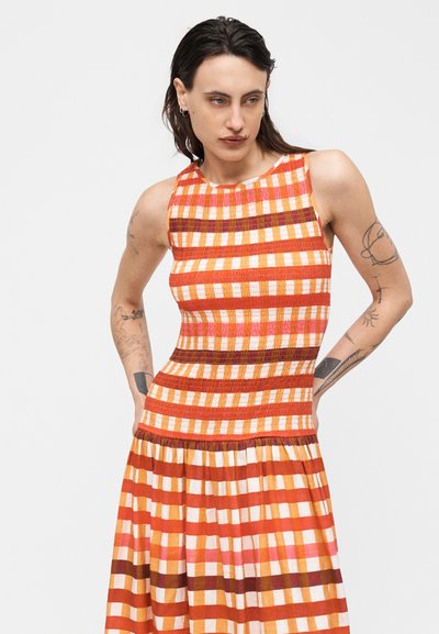 Άτομο με σκούρα μαλλιά μέχρι τους ώμους και τατουάζ, ντυμένο με αμάνικο πορτοκαλί, λευκό και καφέ καρώ φόρεμα, με τα χέρια στη μέση.