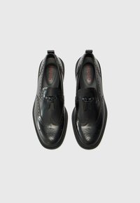 Black leather slip-on shoes featuring brogue detailing, pattern accents, and a textured sole. Interior has "HUGO" branding in red.