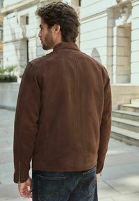 Brūns zamšādas jaka ar paceltu apkakli, ar rāvējslēdzējiem pie piedurknēm un gludu tekstūru, valkāta virs zila džinsu biksēm āra pilsētas vidē.