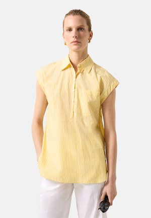 Kobieta ubrana w żółtą, paskowaną koszulkę bez rękawów i białe spodnie, trzyma czarne okulary przeciwsłoneczne w prawej ręce na białym tle.